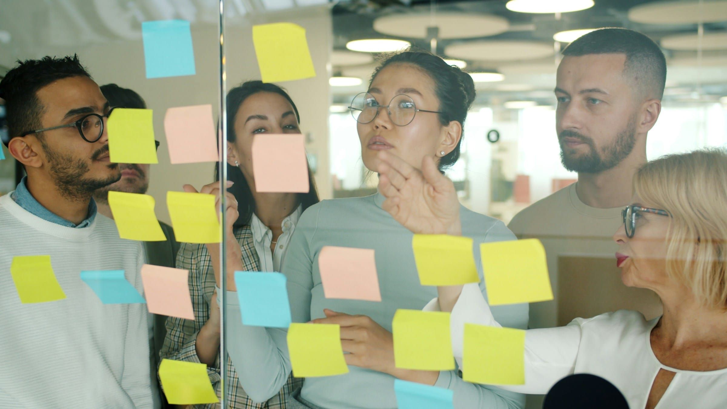 Colleagues gathered at a glass wall covered in yellow, pink and blue sticky notes, sorting ideas in a bright office