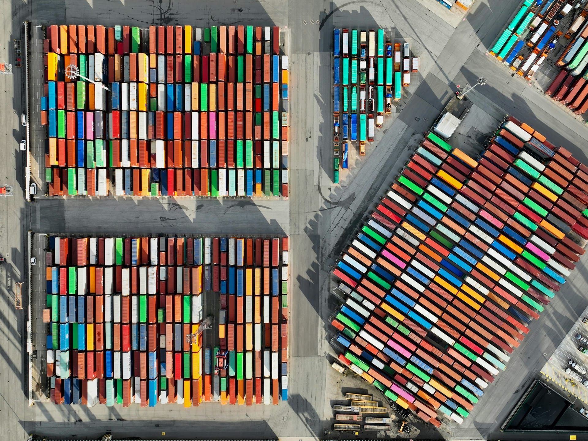 Aerial view of a container port, with rows of red, orange, blue, green and white shipping containers stacked in sunlit yards