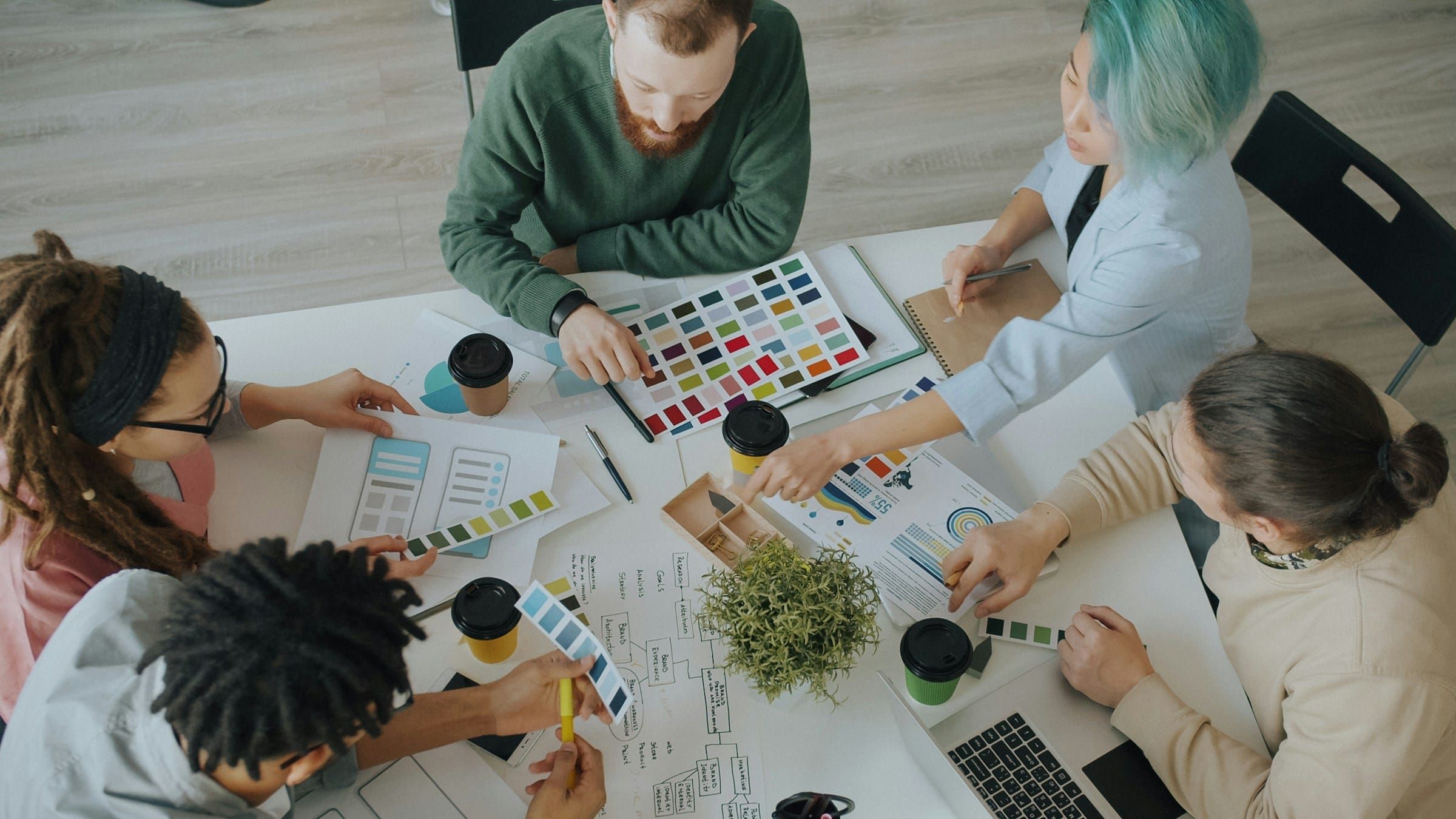 Top-down view of five colleagues around a white table with color swatches, printed charts, a brand plan and coffee cups