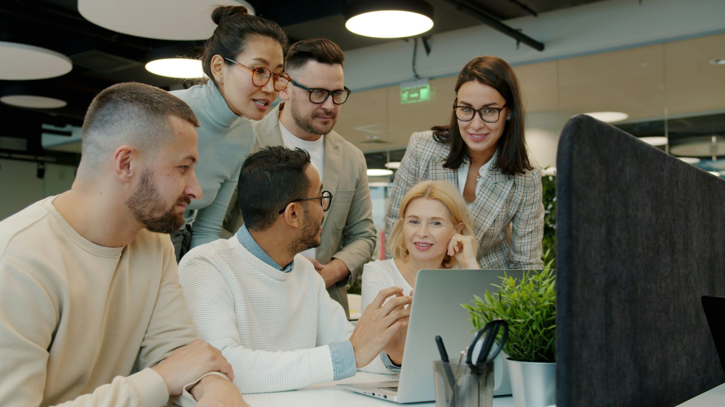 Diverse group of colleagues gathered around a laptop, discussing work in a modern open-plan office