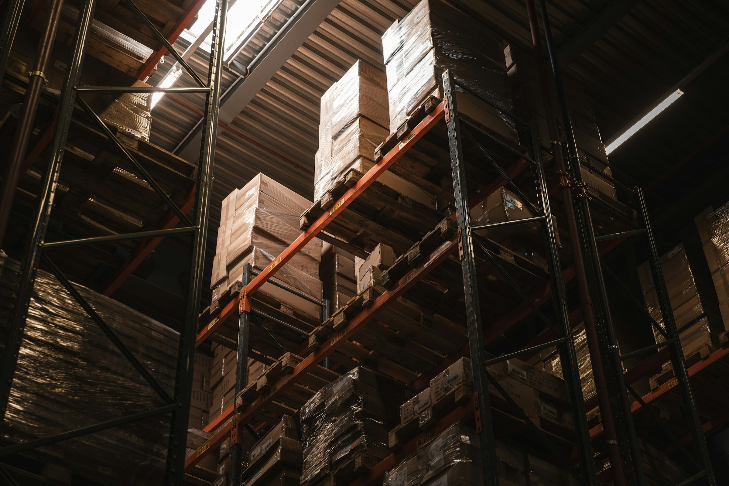 Looking up at tall pallet racking stacked with boxed goods in a dim distribution center