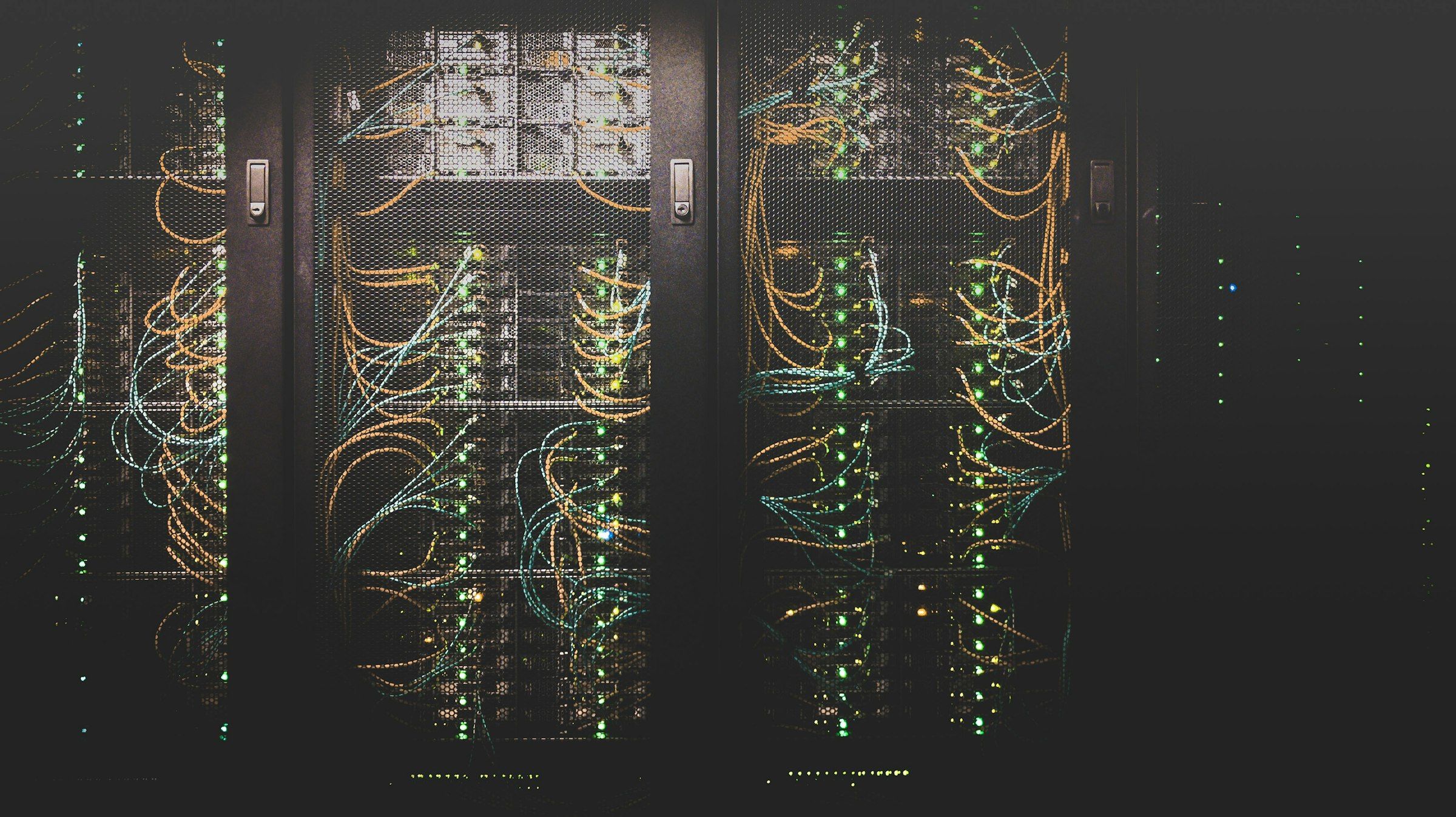 Data center server racks in the dark, behind mesh doors, with green status lights and fiber cabling
