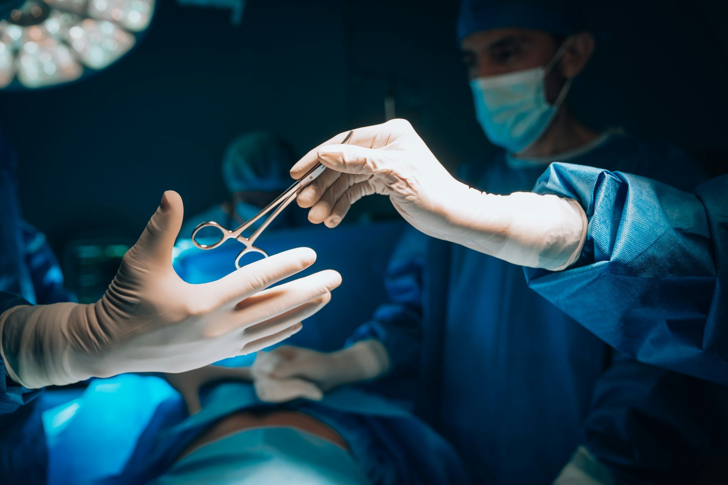 Gloved surgeon passing a surgical clamp to a colleague in a dim, blue-lit operating room