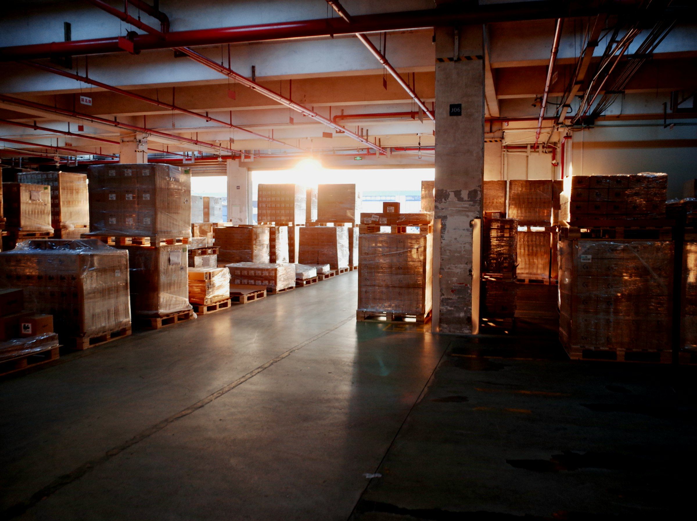 Shrink-wrapped pallets of goods lined up on a warehouse floor, lit by low sun through the loading bay