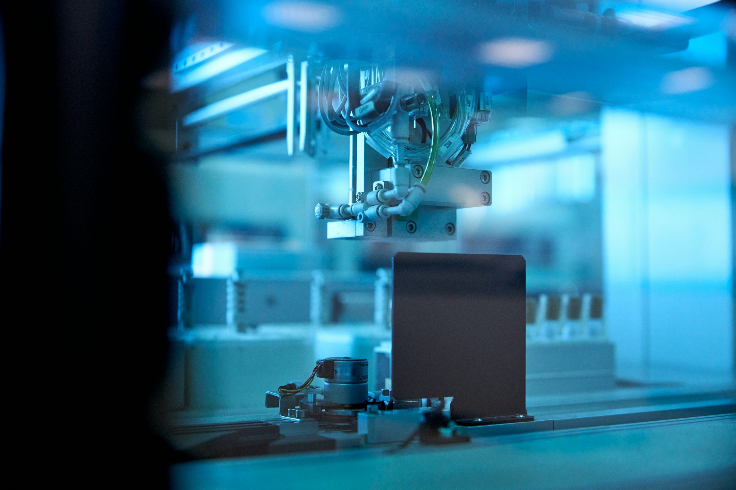 Automated pick-and-place head with pneumatic lines above a part, seen through glass in blue light